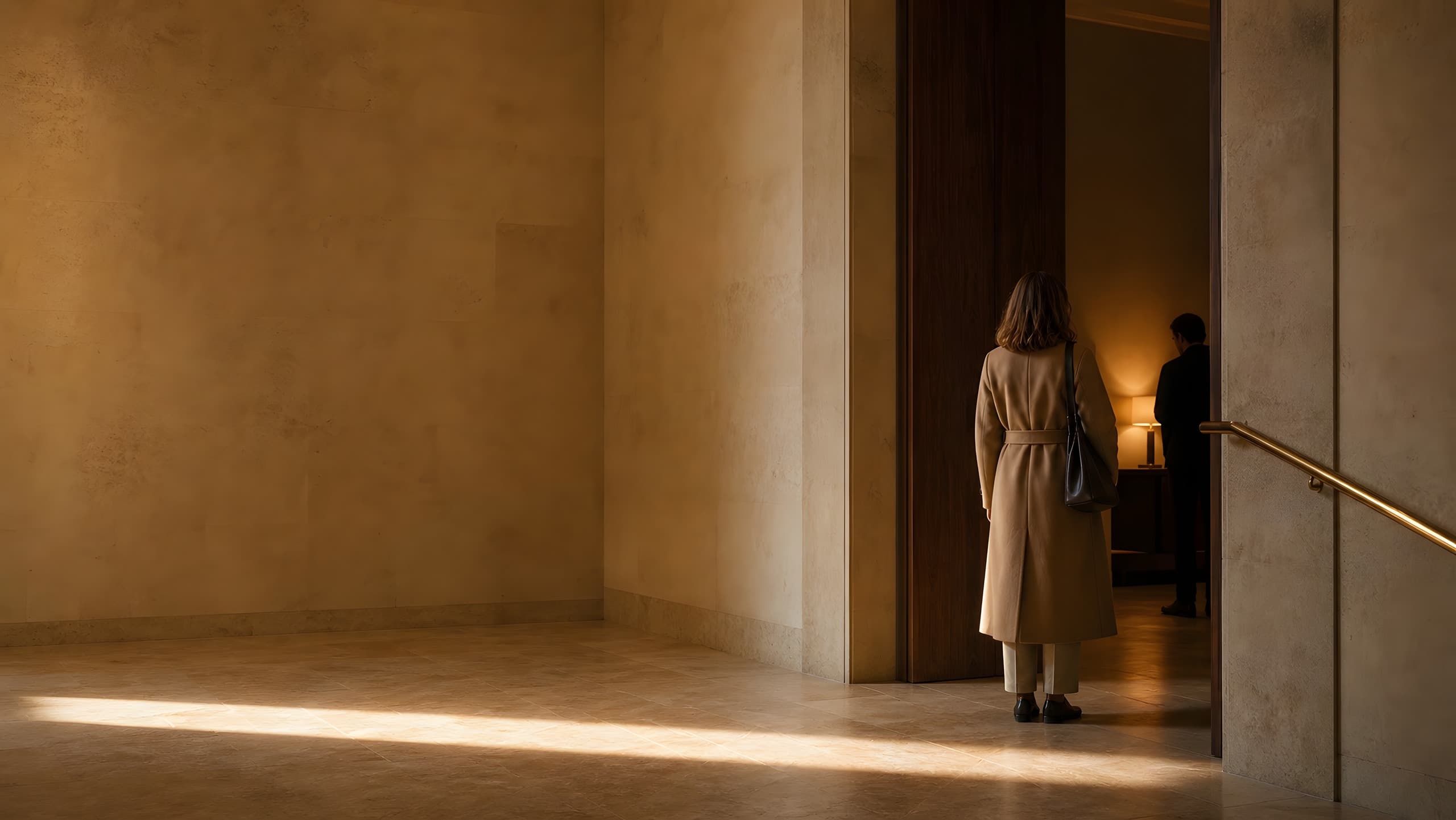 A visitor in a camel coat standing at a tall walnut threshold in a warm limestone hall, late sun across the floor, a lamplit room beyond.